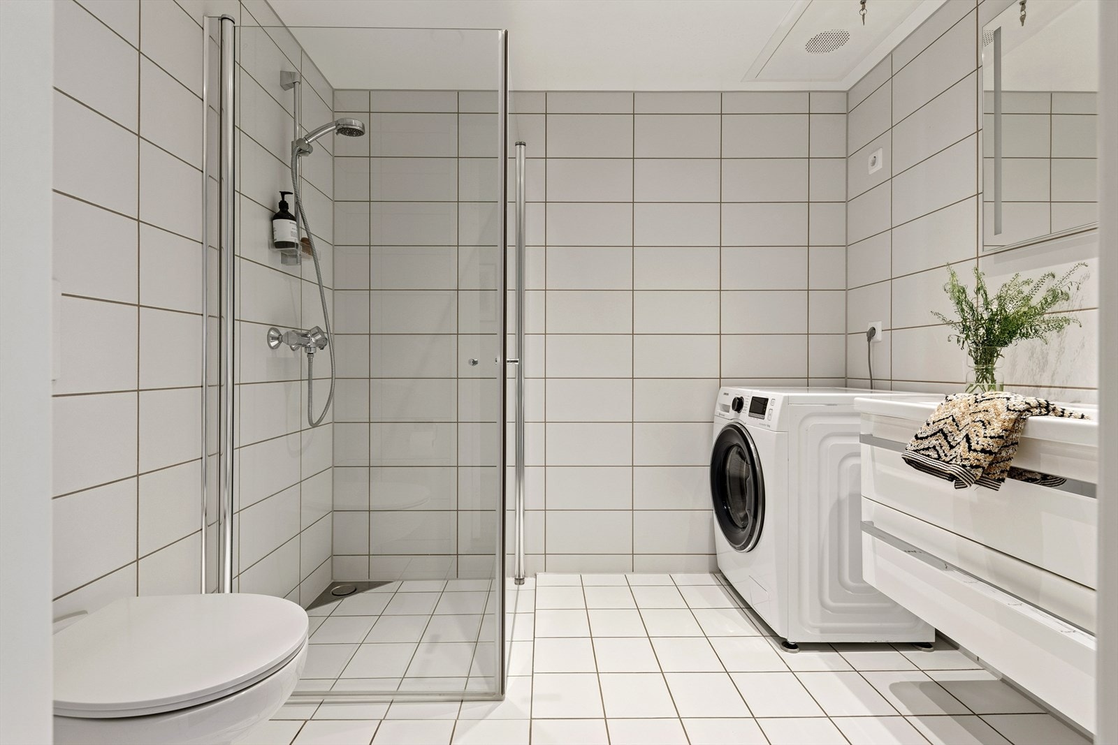 Rommet er utstyrt med vegghengt toalett, hvit baderomsinnredning, speil med integrert belysning, dusjhjørne og opplegg for vaskemaskin Galleribilde