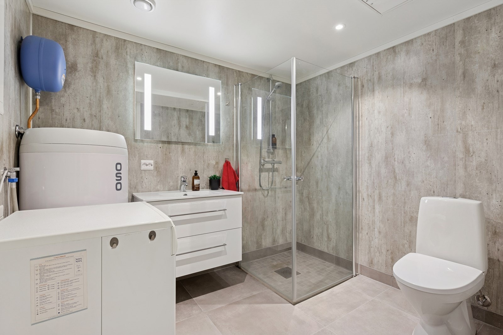 Lyst og pent bad utstyrt med dusjhjørne, gulvstående toalett og opplegg for vaskemaskin. Galleribilde