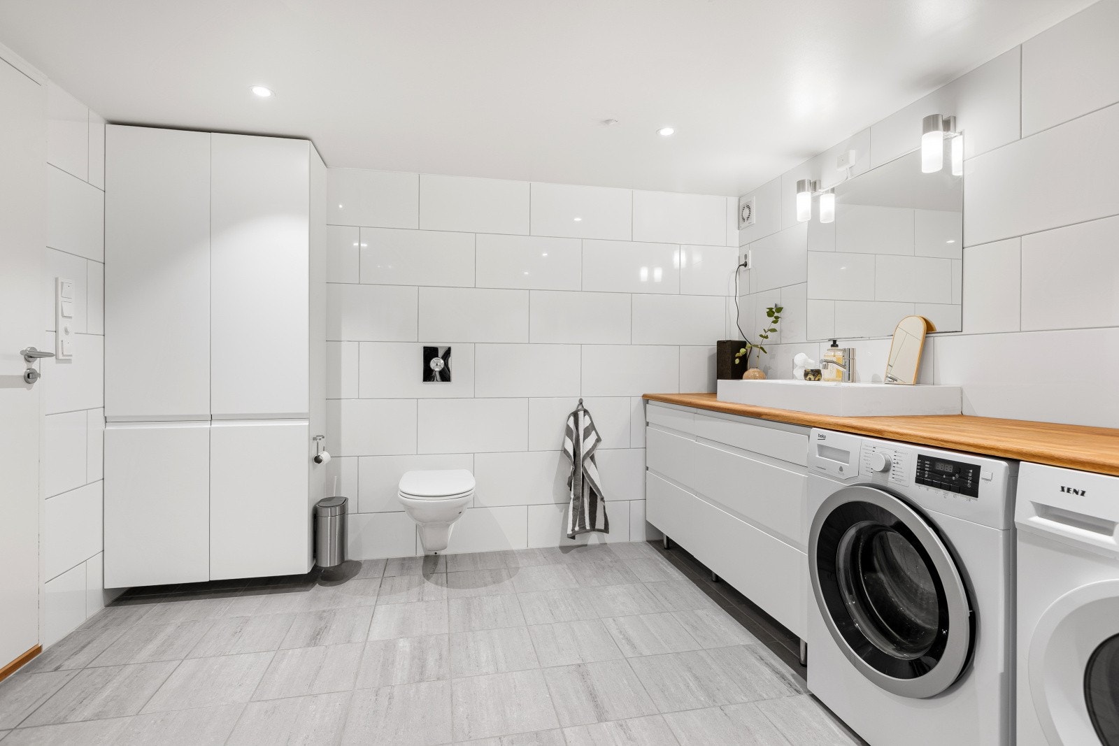 Badet er innredet med en servant med underskap, speil med belysning, dusjvegger og et vegghengt toalett. Det er også opplegg for vaskemaskin. Galleribilde