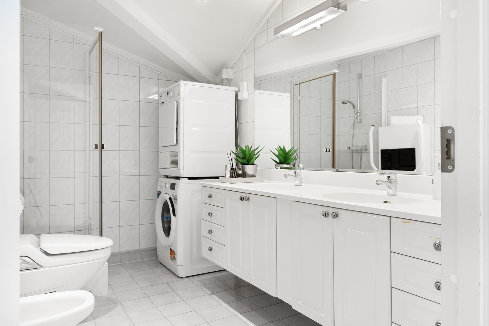 Baderom med fliser på gulv og vegger. Badet er innredet med eldre vegghengt innredning med to servanter og speil på vegg, samt dusjkabinett, elektrisk klosett og bidé. Galleribilde