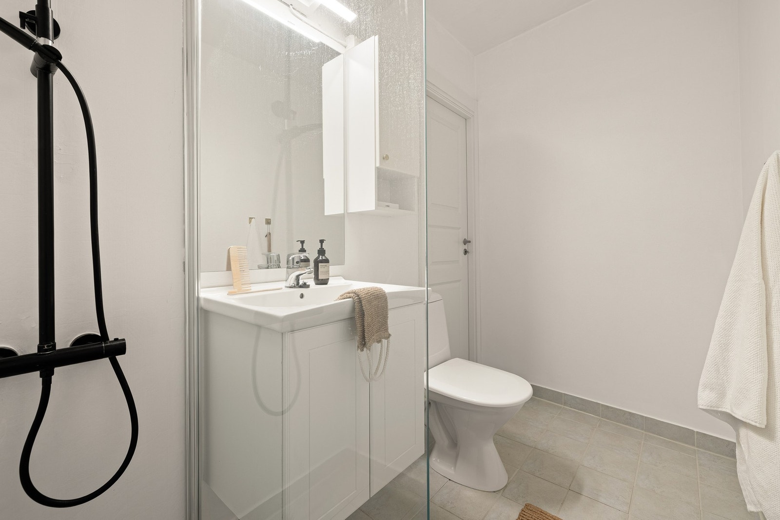 Badet er innredet med toalett, dusj med glassvegg og sort dusjgarnityr, og baderomsinnredning med speil og belysning. Galleribilde