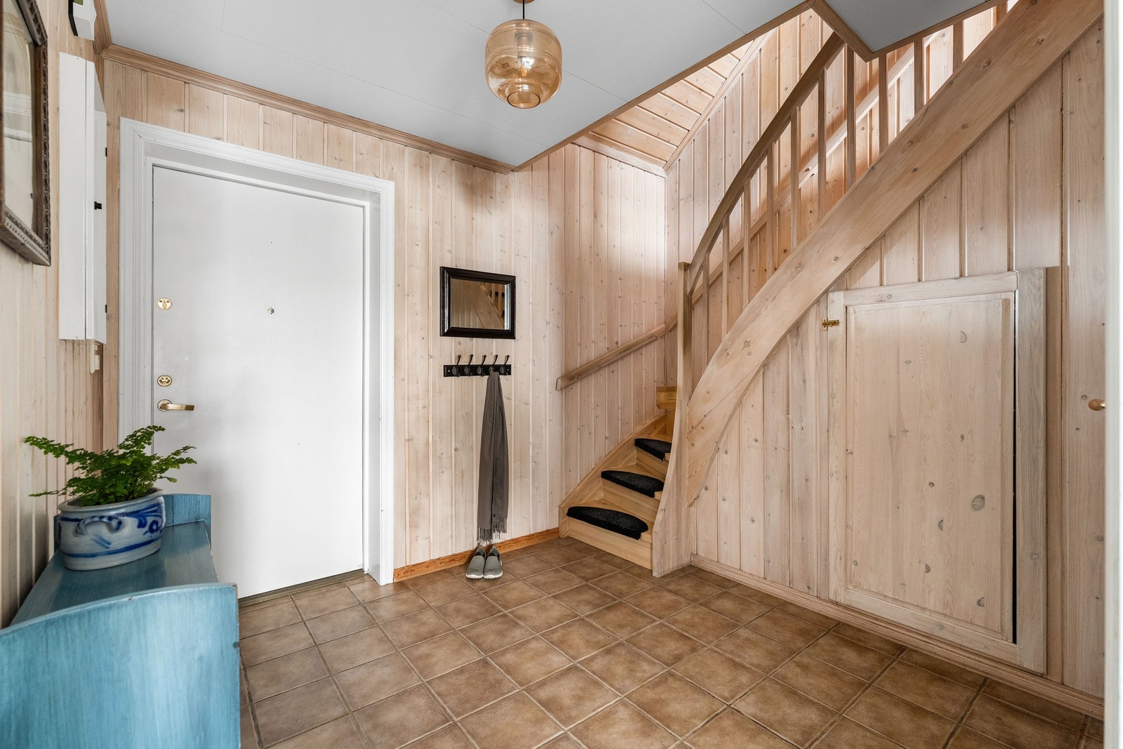 Tiltalende entré med fliser på gulv med gulvvarme og kalkbehandlet panel på veggflater som gir et godt førsteinntrykk. Det er bygget inn en praktisk skapløsning under trappen for ekstra lagring. Galleribilde