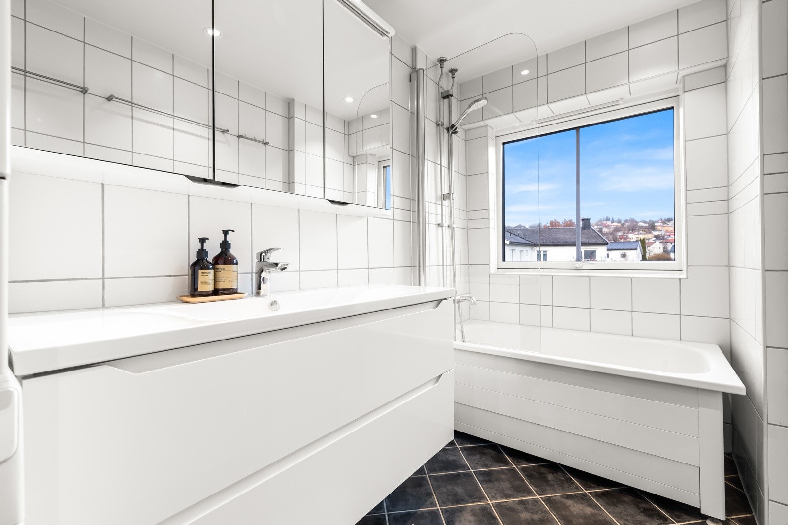 Badet har stilige fliser på vegger og gulv med downlights i himling. Det er innredet med innredning med skuffer, heldekkende servant med ettgreps blandebatteri, veggmontert toalett, badekar med dusjvegg på hengsler. Galleribilde