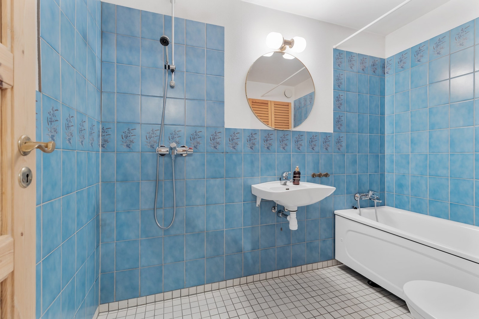 Bad fra byggeår. Flislagt gulv med gulvvarme. Flislagte vegger samt malt strietapet. Slett malt himling. Vegghengt servant med ett-greps armatur. Speil og overlys på vegg over servant. Badekar i hjørne. Blandebatteri tilkoblet hånddusj. Dusj på gulv. Galleribilde