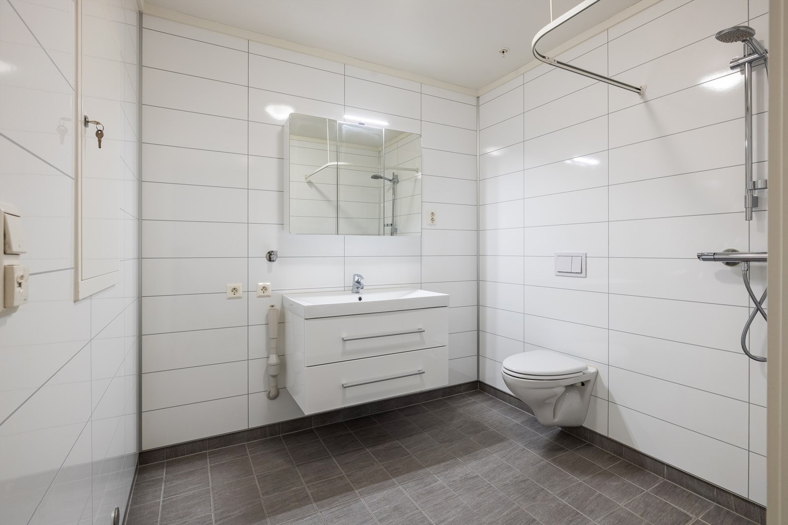 Flislagt baderomsgulv og tidløse baderomsplater på vegger. Dusjhjørne, vegghengt toalett, servant og opplegg til vaskemaskin/tørketrommel Galleribilde