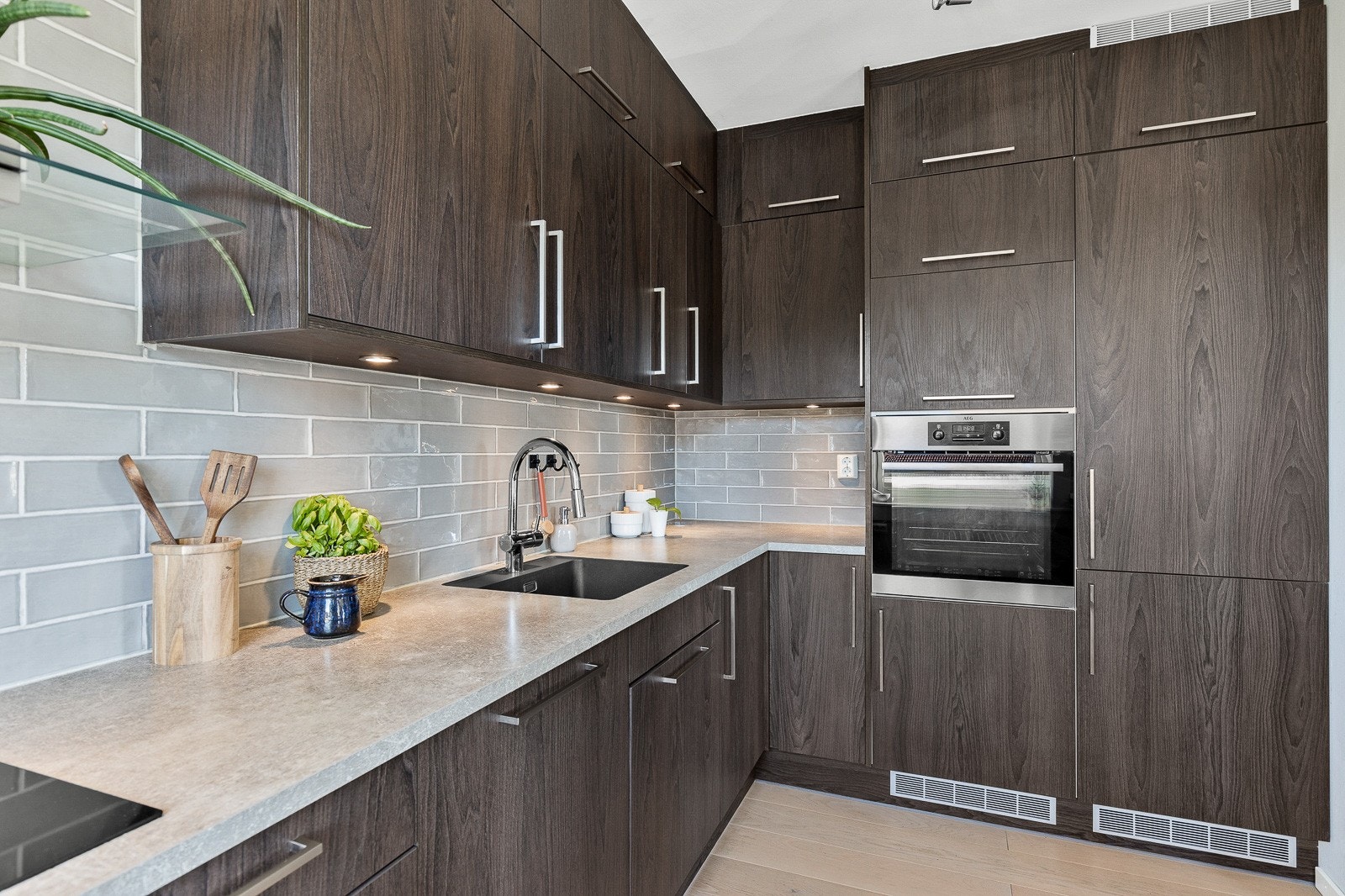 Kjøkkeninnredning med glatte fronter, laminat benkeplate med nedfelt kum i rustfritt stål, fliser på vegg og belysning over benkeplaten. Galleribilde