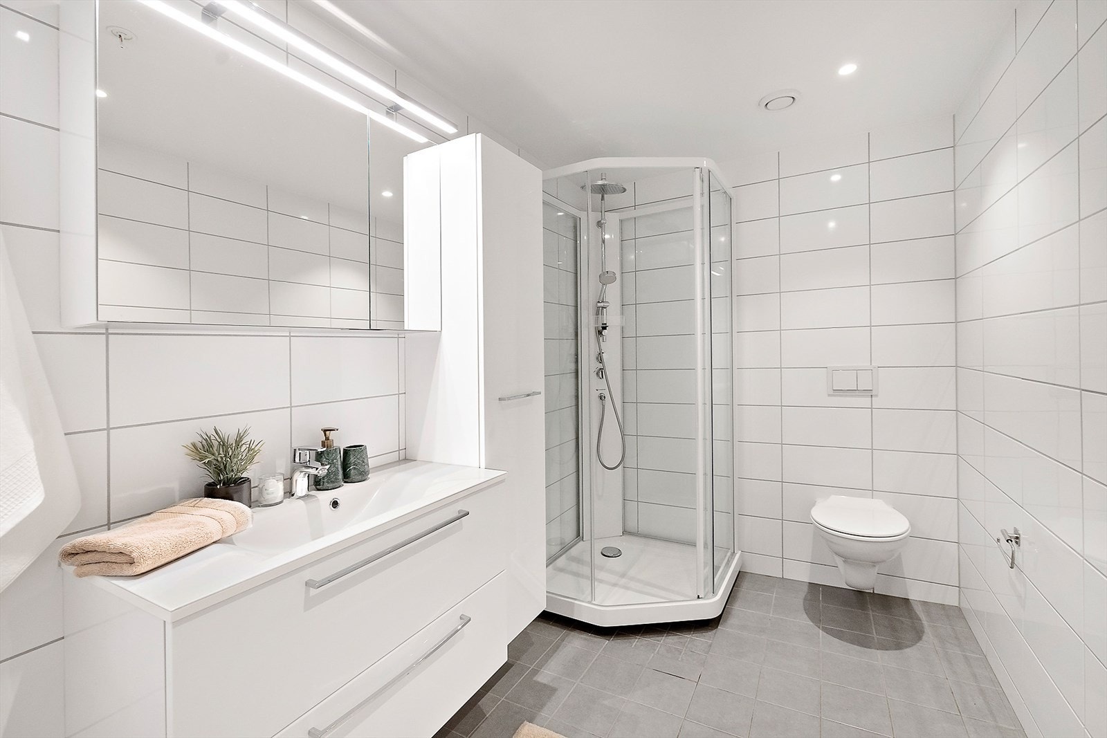 Pent bad med hvit innredning. Servant med skuffer, speil med integrert lys, dusjkabinett og wc. Galleribilde