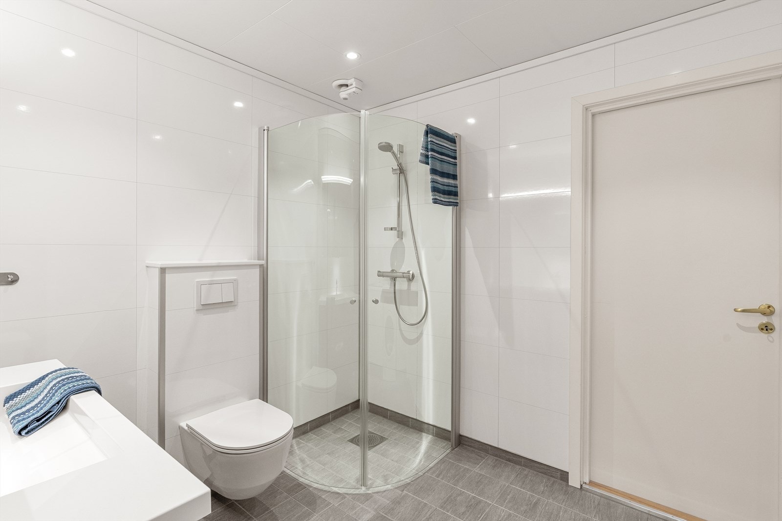 Badet er innredet med servant i skuffeseksjon med overhengende speilskap, vegghengt toalett og dusj med glassvegger som kan slås inn. Galleribilde