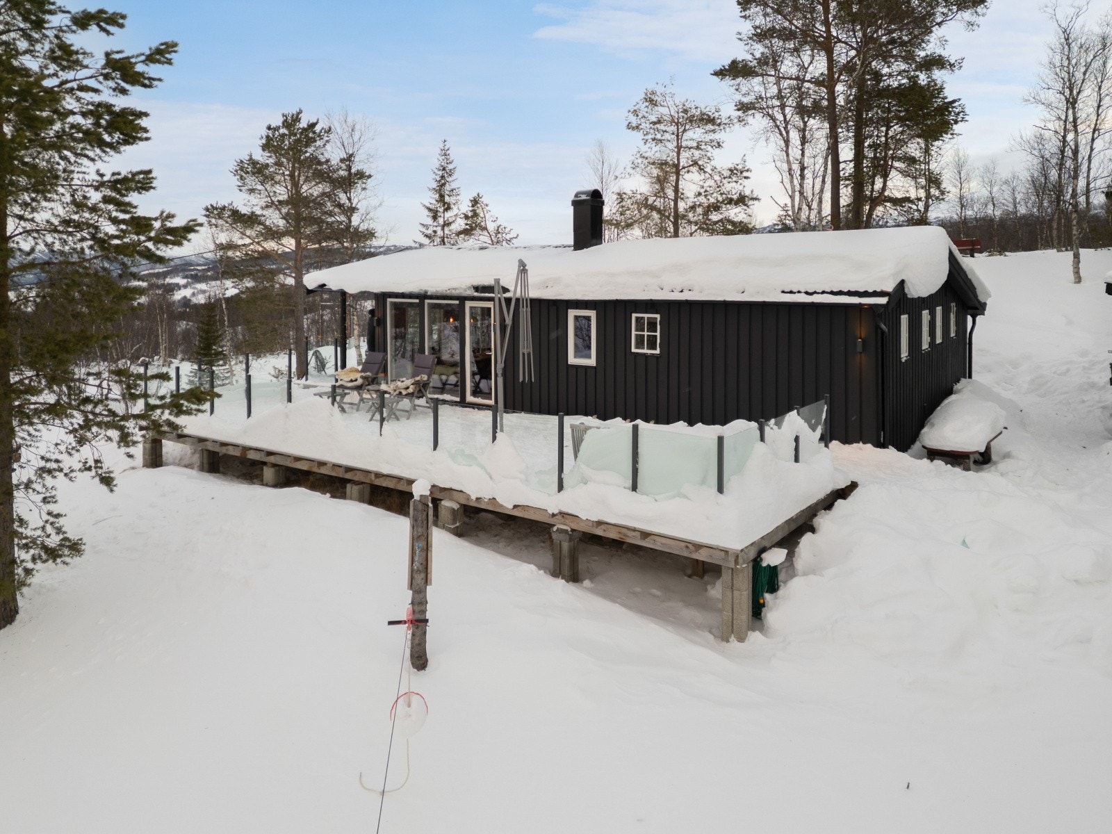 Fritidsboligen har en terrasse i impregnert trevirke med glassrekkverk, plassert på pilarer. Galleribilde