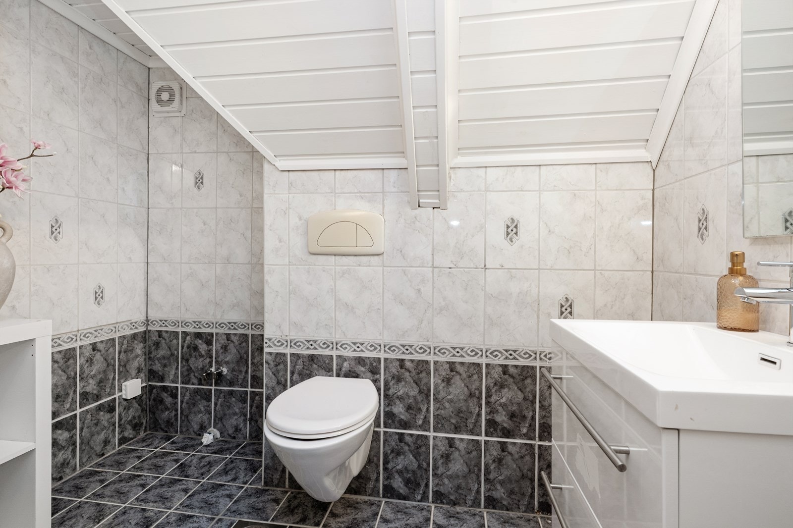 Flislagt toalettrom som er innredet med vegghengt toalett og servantmøbel med skuffer og speil. Galleribilde