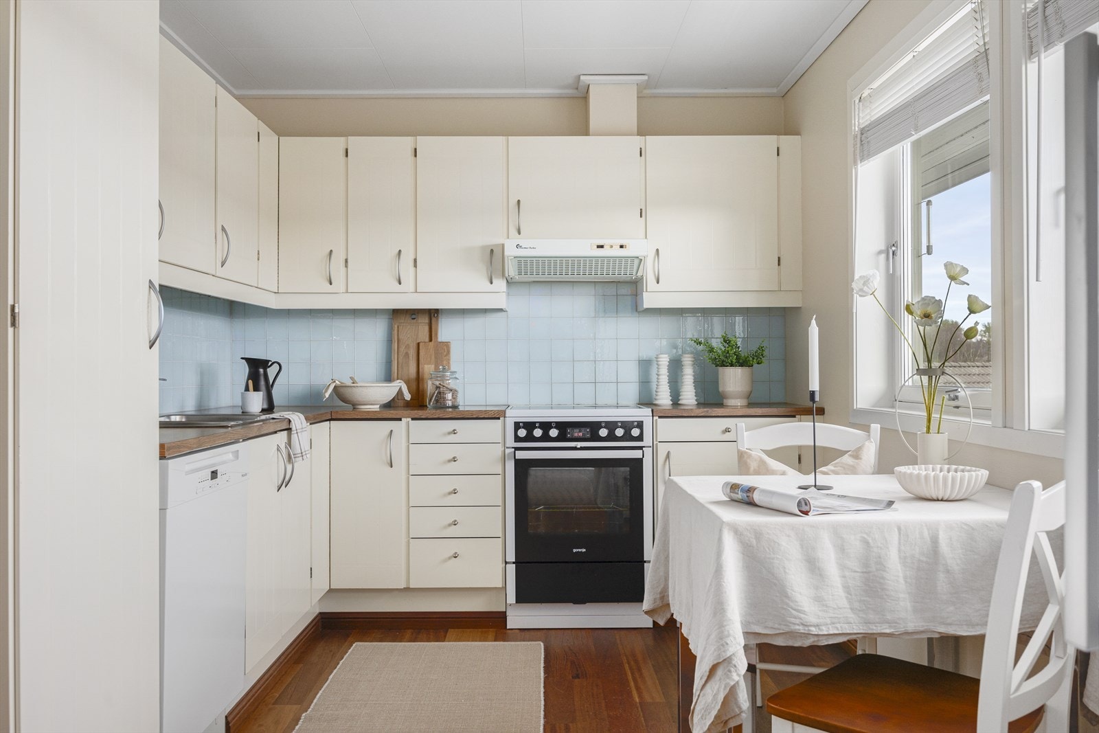 Kjøkkeninnredning med lyse fronter og benkeplate i laminat med nedfelt vask. Glassflisene på veggen gir et moderne preg, og den åpne løsningen gir plass til lite spisebord. Galleribilde