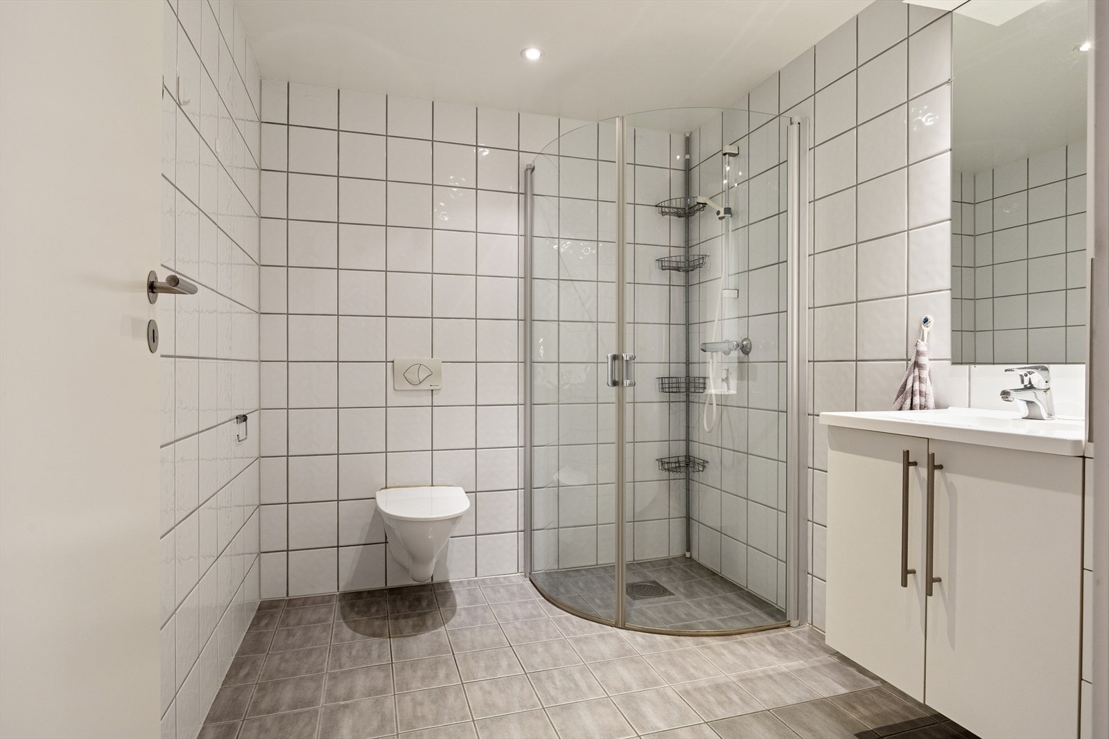 Baderommet er tidløst innredet med grå fliser på gulvet og hvite fliser på vegg. I gulvet har man også varmekabler for ekstra komfort. Badet er ellers utstyrt med dusjhjørne, vegghengt toalett og baderomsinnredning med god lagringsplass og nedfelt servant Galleribilde