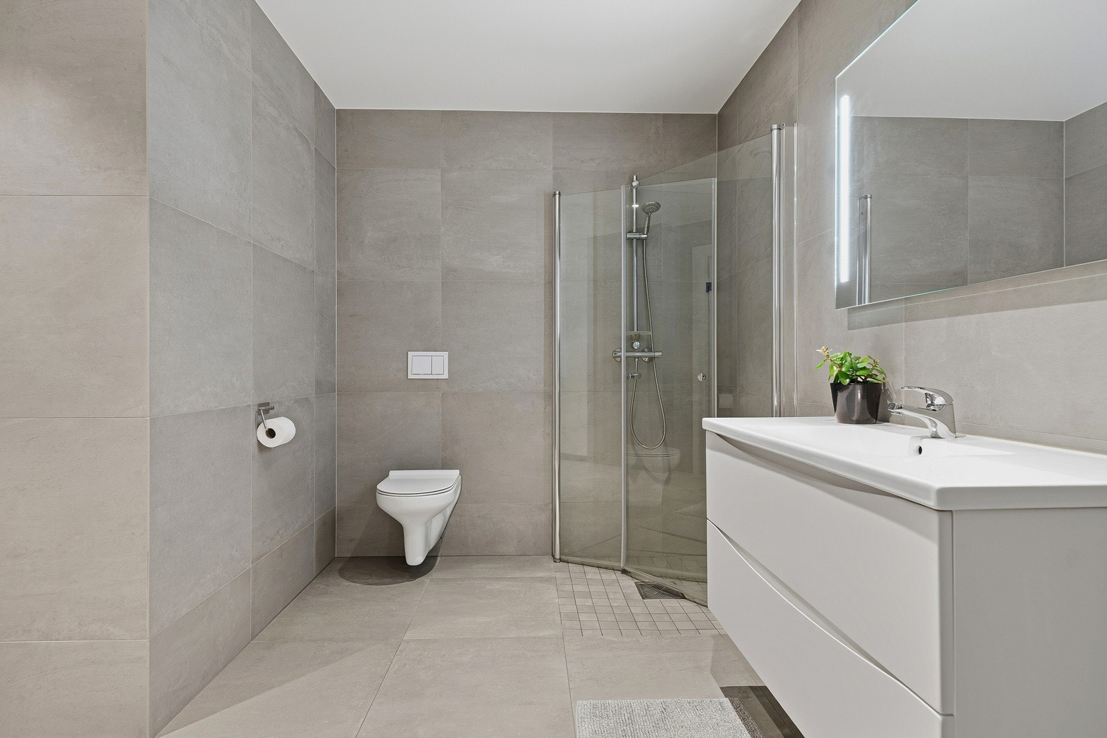 Lekkert baderom bestående av vegghengt toalett, baderomsinnredning og dusjhørne med innfellbare dusjdører. Galleribilde