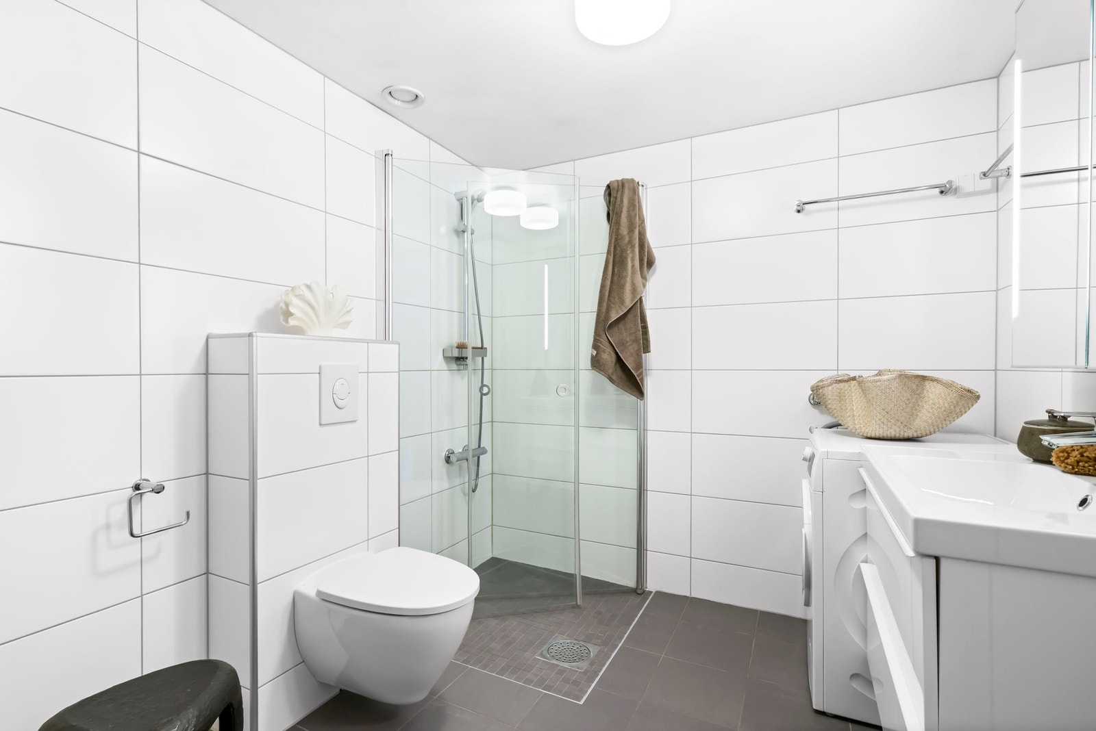 Stort lyst baderom med opplegg for vaskesøyle for vaskemaskin og tørketrommel, praktiske innslagbare glassdører i dusjsonen i herdet klart glass og vegghengt toalett. Galleribilde