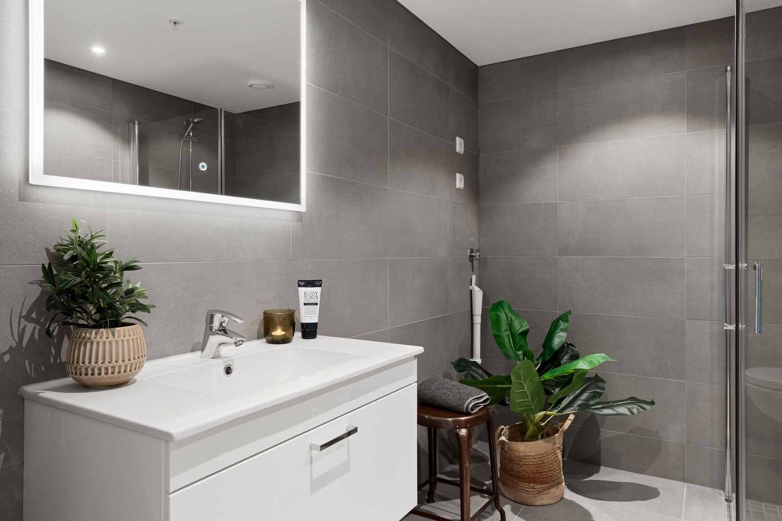 Moderne bad med grå fliser, dusjkabinett, hvit servant og speil med lys. Galleribilde
