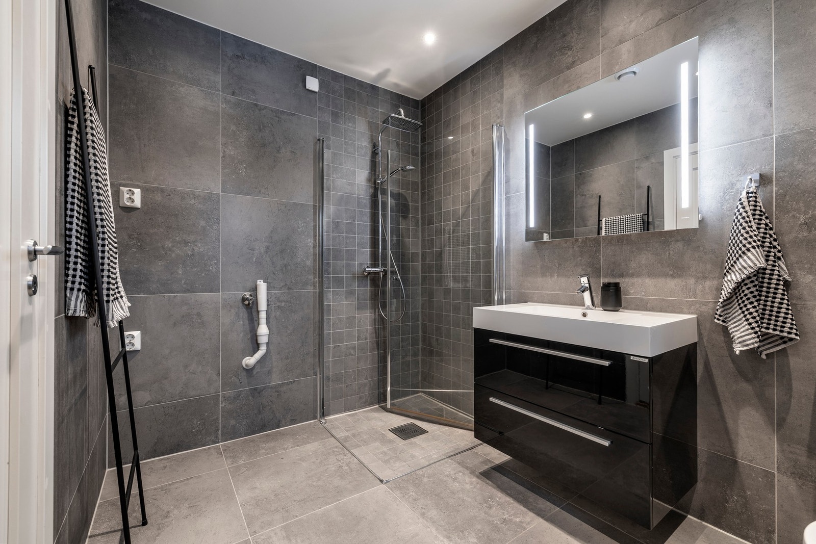 Badet er innredet med servant, speil med lys på vegg over servant, vegghengt toalett og dusjhjørne med svingbare glassdører. Galleribilde