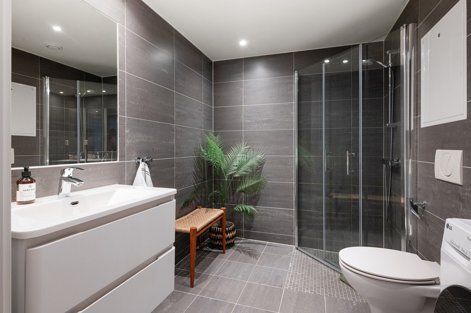 Romslig og moderne bad med vegghengt toalett, servant nedfelt i innredning, speil og dusjhørne med svingbare glassdører. Galleribilde