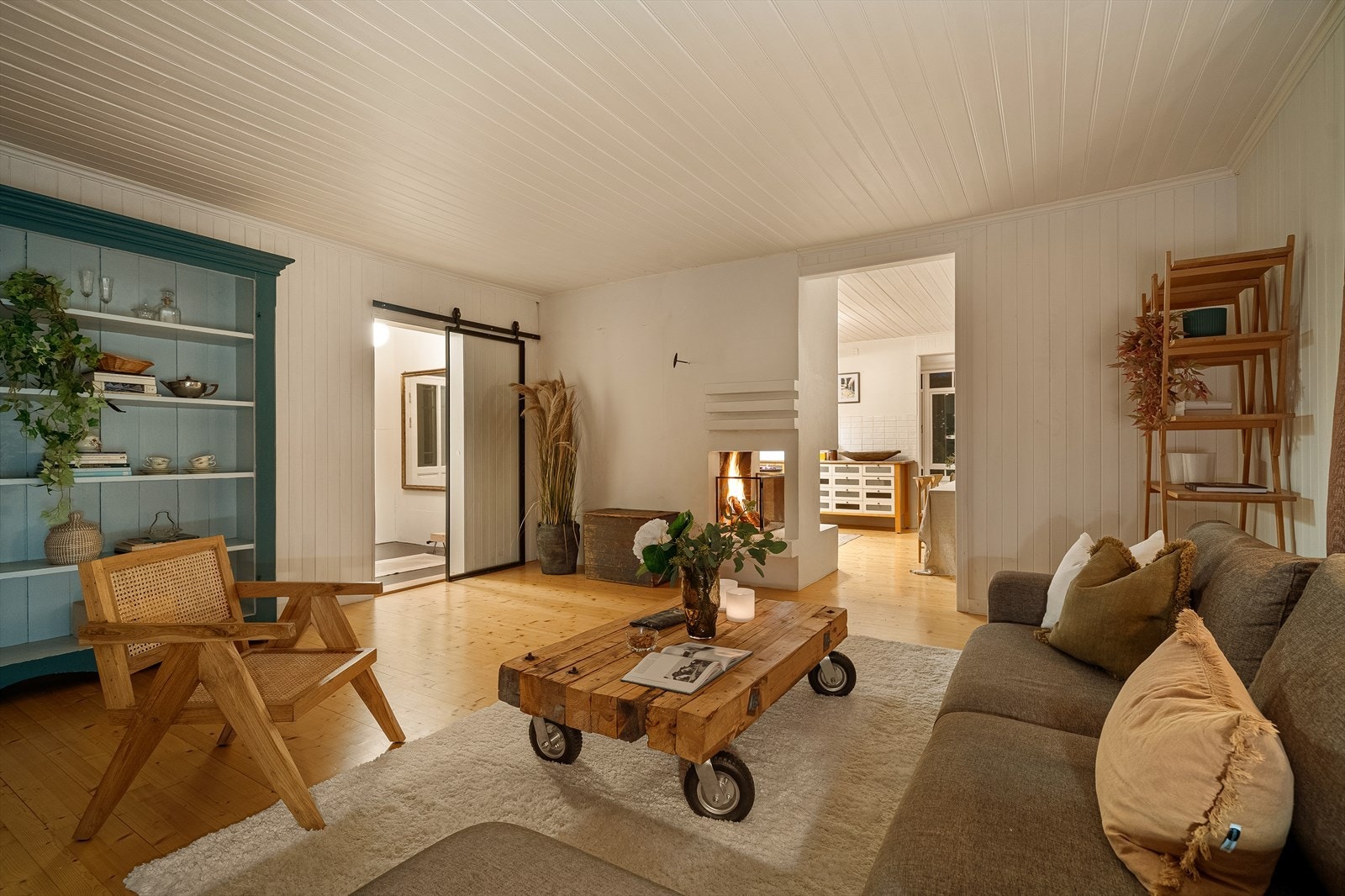 Hovedhusets stue byr på en behagelig overgang til kjøkkenet, og den koselige peisen skaper en innbydende ramme for hverdagsliv og gjester Galleribilde