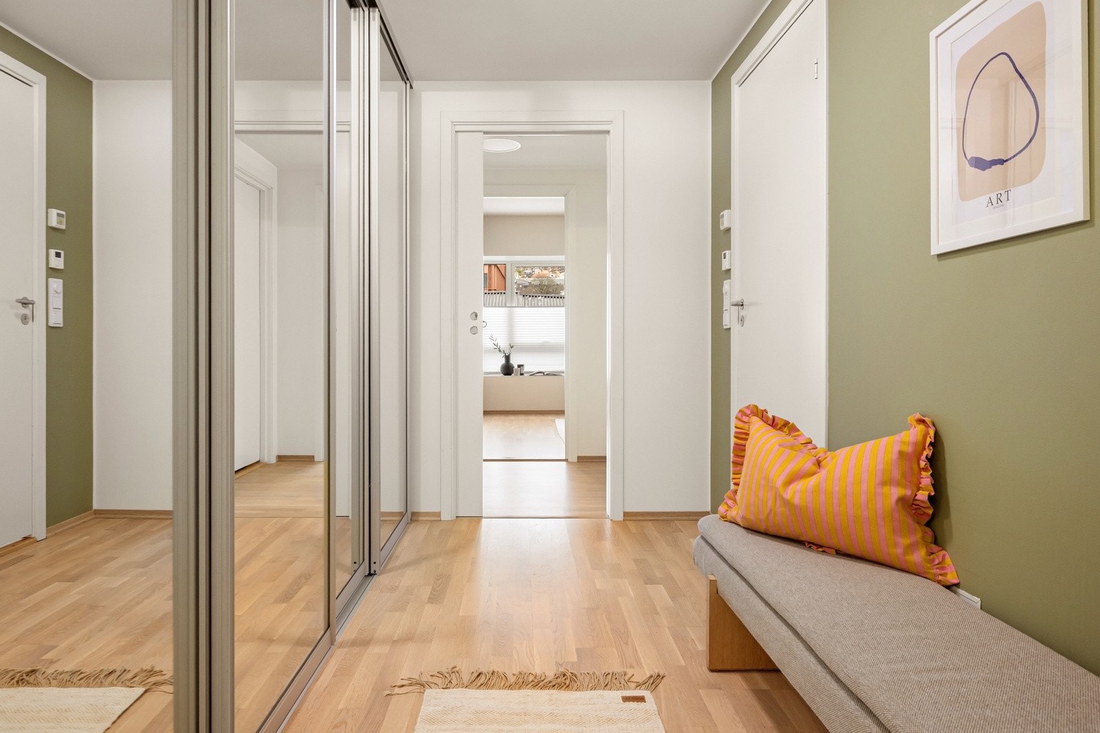 Entréen er romslig og måler 6,4 m². Den er utført med lys eikeparkett og tilpassede gulvlister, og byr på god plass til yttertøy i praktisk skyvedørsgarderobe med speilfronter. Dørtelefon med portåpner er plassert lett tilgjengelig ved inngangsdøren. Galleribilde