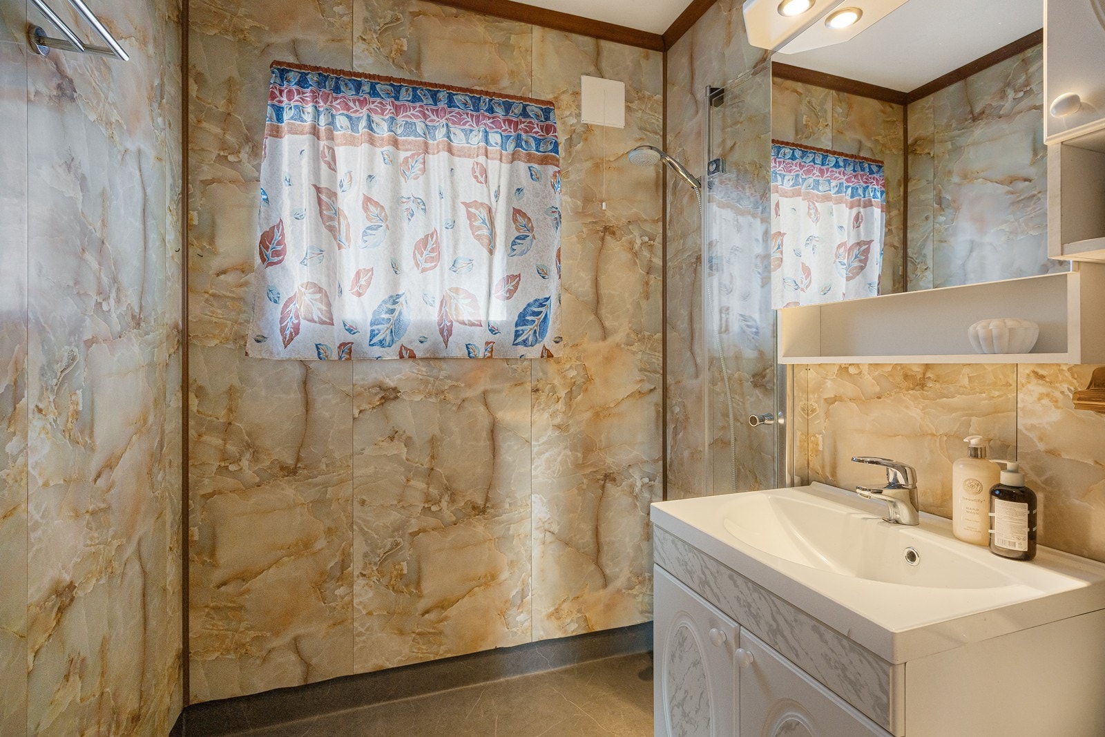 På badet er det toalett og et åpent dusjhjørne. Den øvrige innredningen består av et servantskap med profilerte fronter, et lite veggskap i samme stil og et speilskap med tilhørende lys. Galleribilde