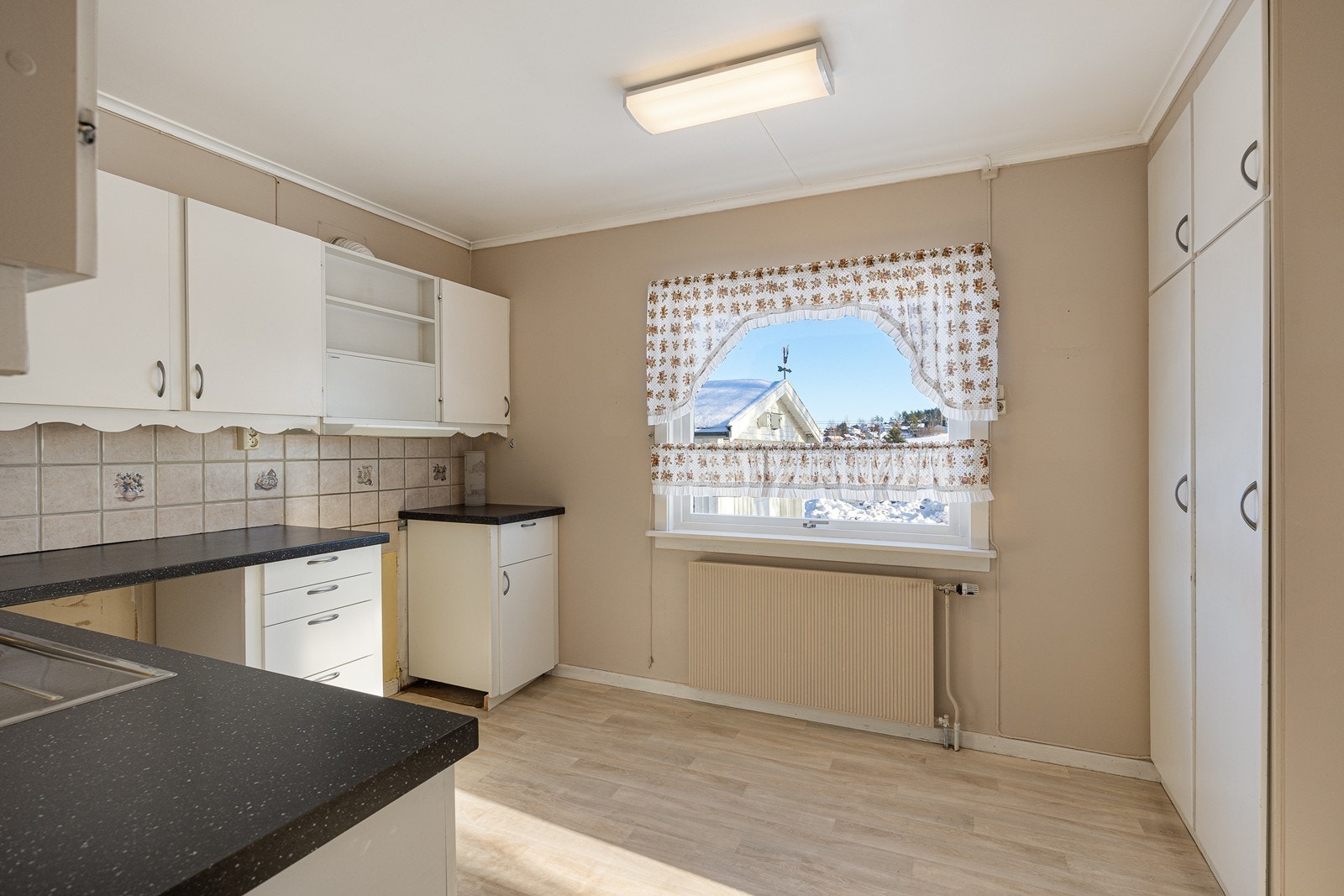 Innredningen har hvite, glatte fronter, benkeplate av laminat og dobbel oppvaskkum i stål. Over benken er det belysning og lysegrå fliser med noe dekor, og over kokesonen er en ventilator etablert med avtrekk ut. Galleribilde