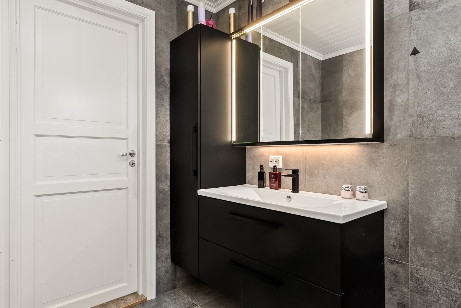 Servantinnredning med mørktonede fronter og speil med belysning gir praktisk oppbevaring. Galleribilde