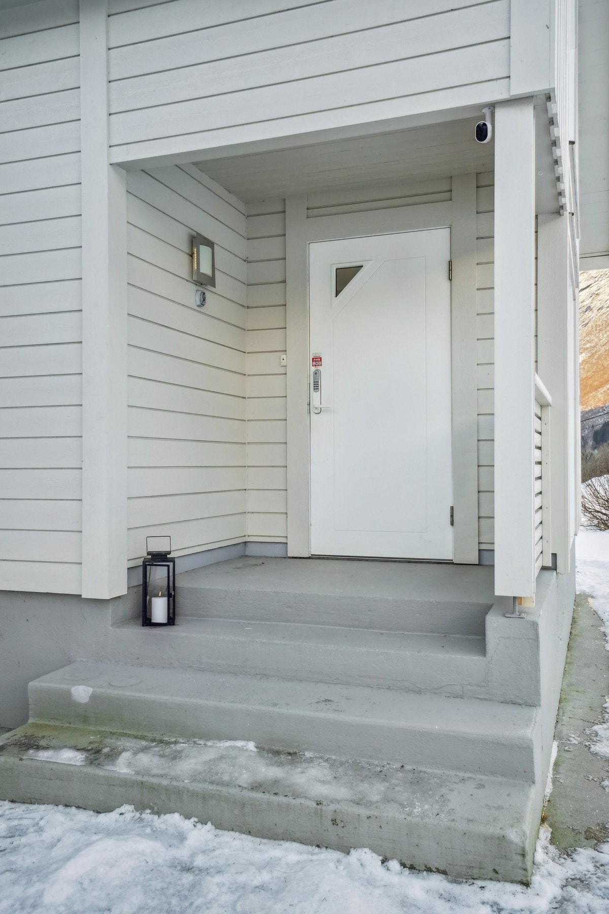 Pent opparbeidet inngangsparti med trappetilkomst og overbygg. Det er montert utebelysning på vegg. Montert kodelås på ytterdøren. Galleribilde