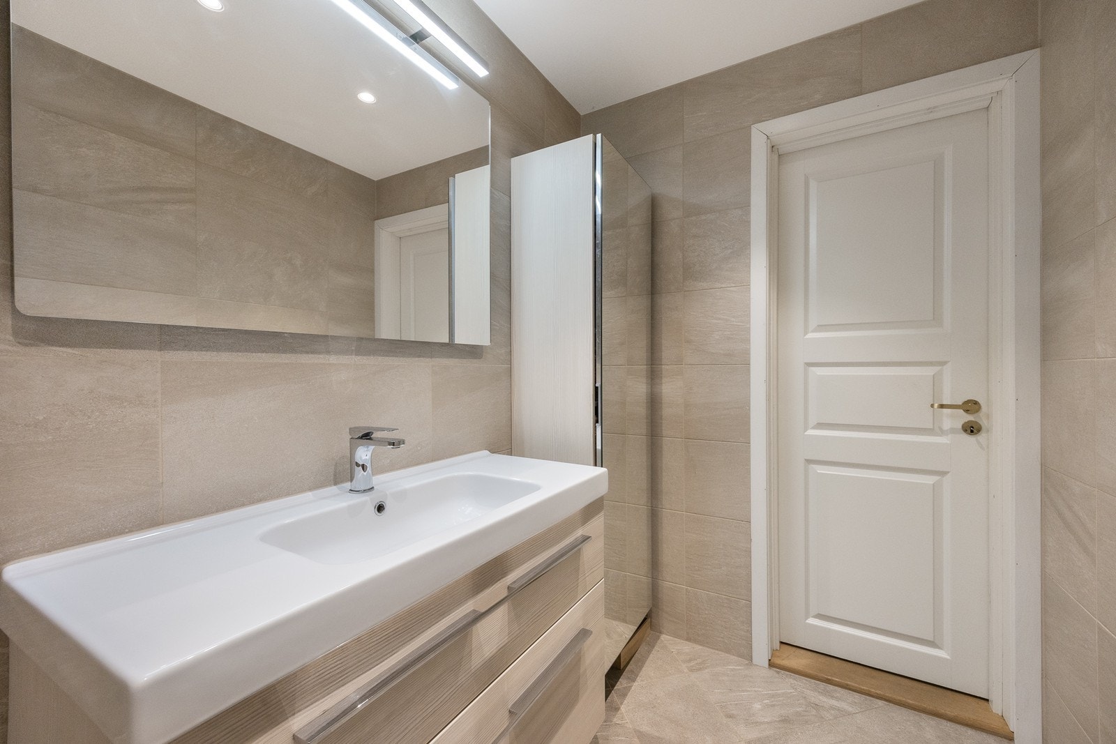 Badet ble pusset opp i 2015, innredet med store, grå vegg- og gulvfliser for et delikat uttrykk. Innredningen består av en servant med lyse skuffer, et høyskap med speilfront og et stort speil med belysning. Galleribilde