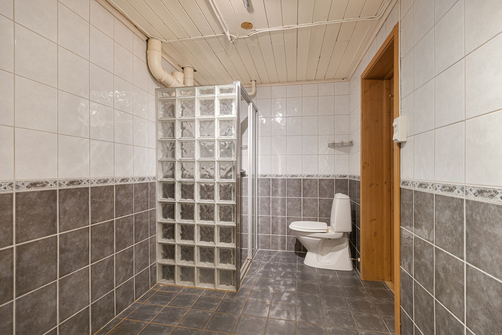 Kjellerbadet har hvite og grå fliser. Det er toalett og gulvvarme, samt et dusjhjørne med skyvedør og vegg av glassbyggerstein. Vaskerommet ligger innenfor badet, innredet med utslagsvask og varme i gulvet. Galleribilde