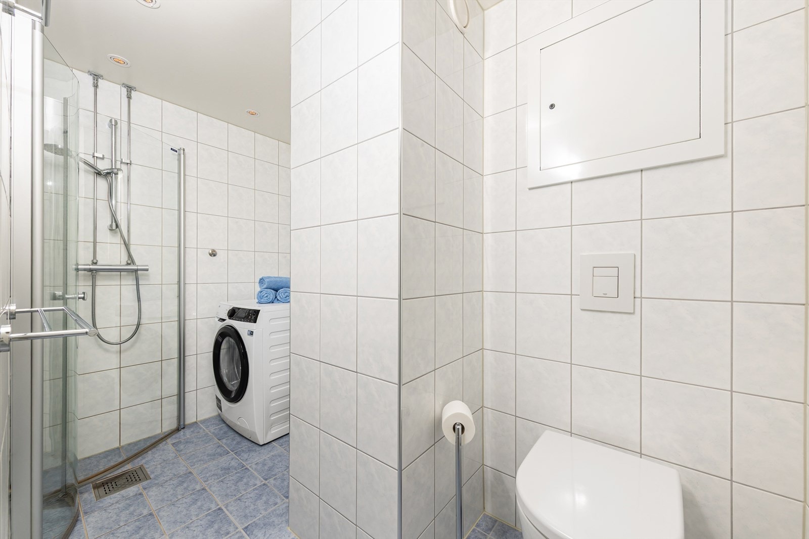 Innredningen består av nedfelt servant, veggmontert toalett, dusjhjørne med glassvegger og praktisk opplegg for vaskemaskin. Galleribilde