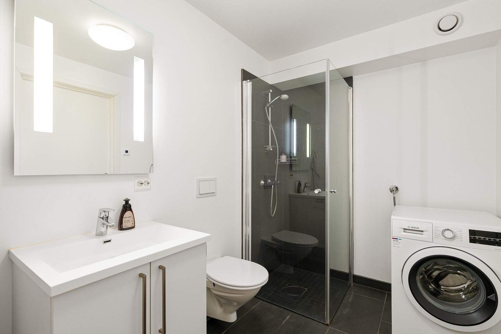 Badet er moderne og praktisk utført med fliser på gulv og i dusjsone, vegghengt toalett, servant i seksjon og speil med integrert belysning. Det er varmekabler i gulv og opplegg for vaskemaskin. Galleribilde