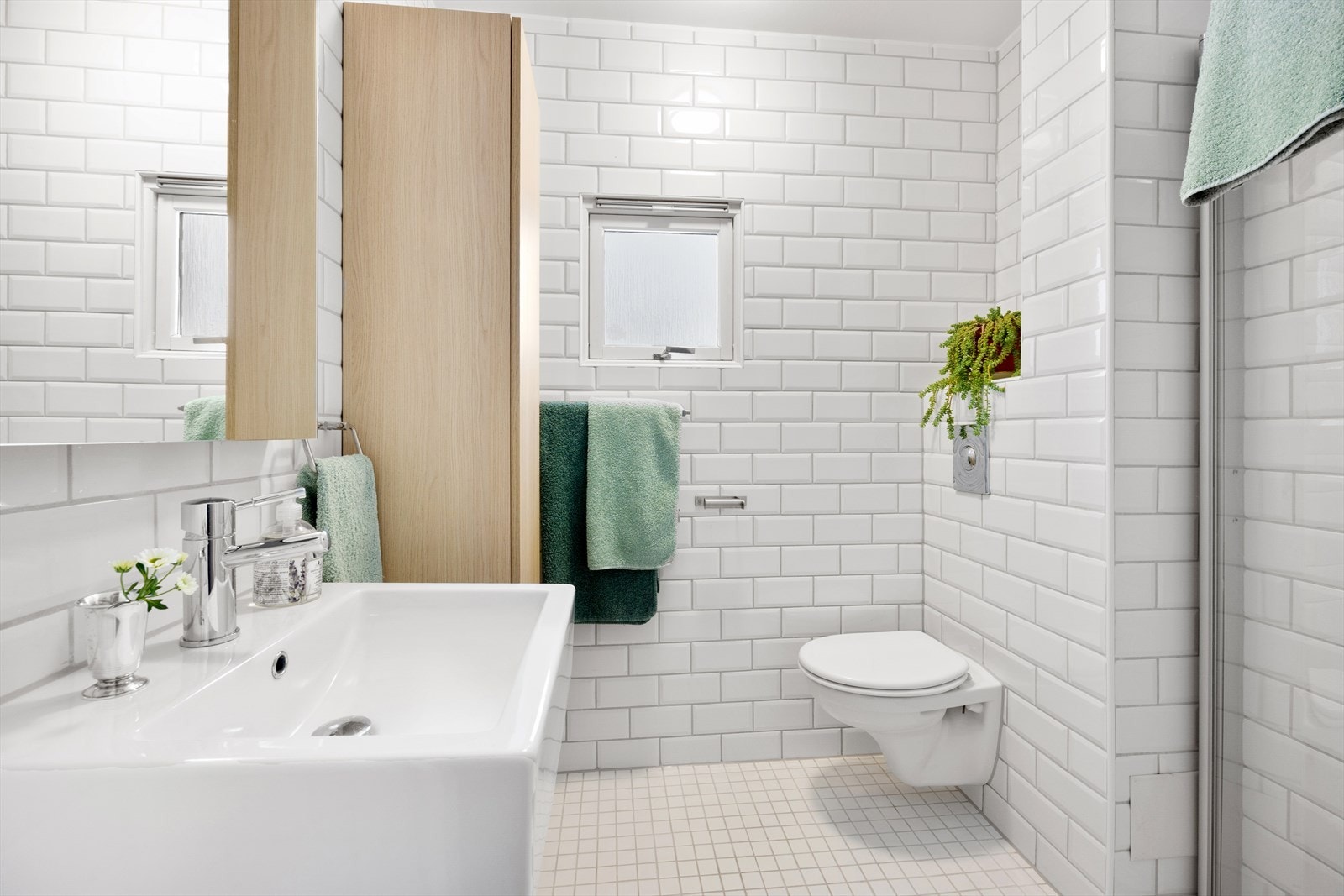 Badet er lyst og praktisk med fliser på gulvet, hvite fliser på veggene, gulvvarme og dagslys fra et lite vindu mot øst. Skapinnredningen er ny fra 2025. Dusj med innfellbare dører i herdet glass, veggfestet klosett med innebygget sisterne, servant og opp Galleribilde