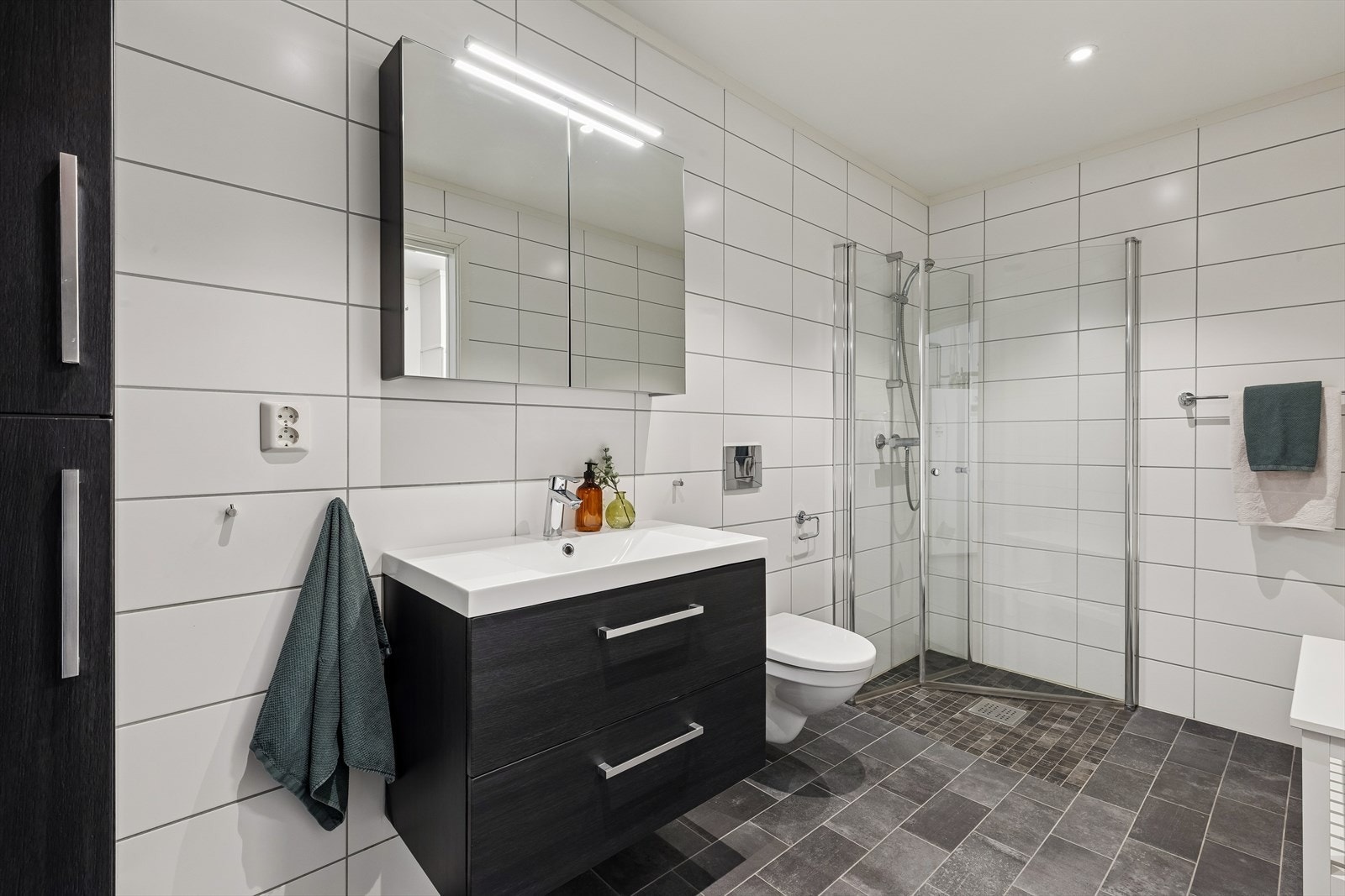 Badet i 1 etasje har fliser på gulv og vegger, veggmontert toalett og dusj med glassdører Galleribilde