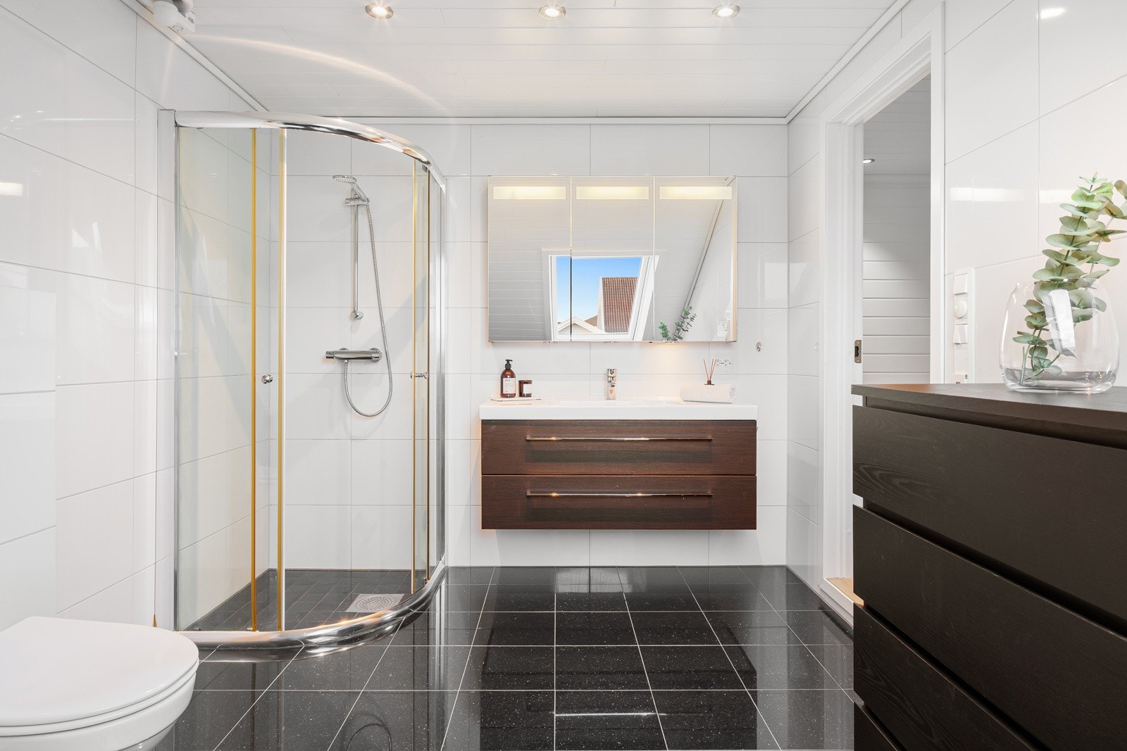 Rommet er innredet med servant på skuffeseksjon og speil med belysning. Videre er rommet utstyrt med toalett, dusjdører og opplegg for badekar. Galleribilde