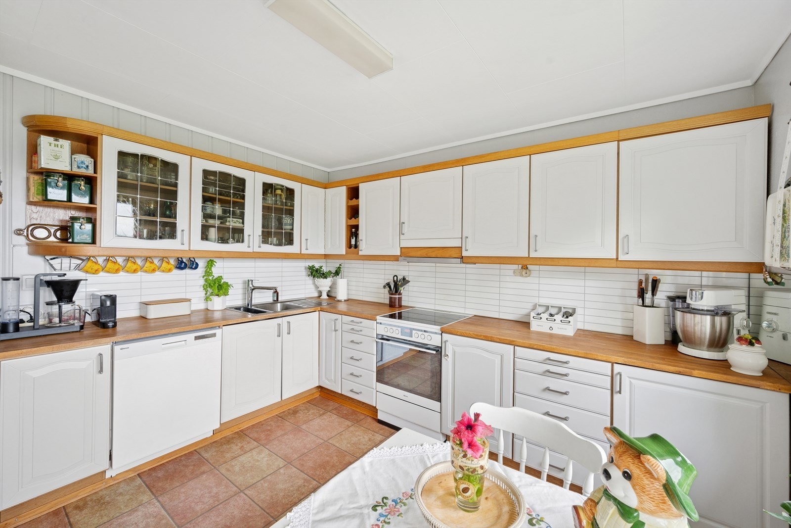 Kjøkkenets innredning har profilerte fronter, belysning under overskap, laminatplate på vegg over benkeskap, heltre benkeplate med nedfelt kum i rustfritt stål og opplegg for oppvaskmaskin. Galleribilde