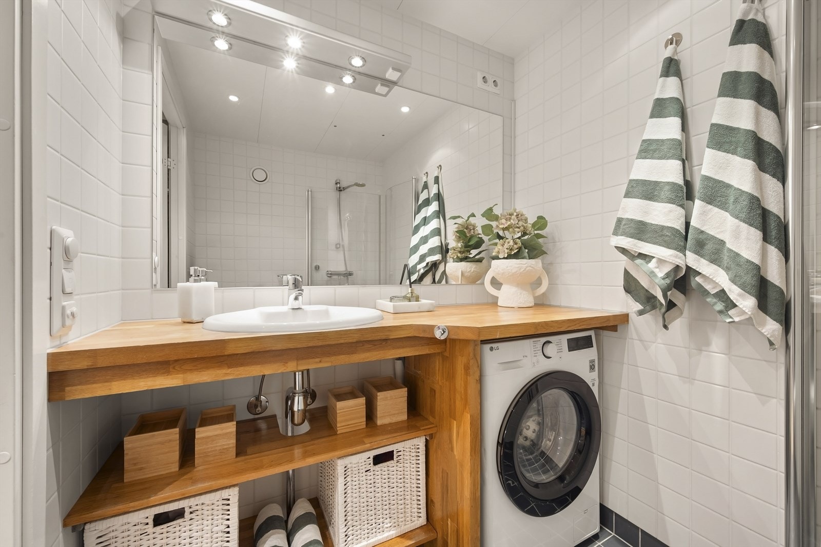 Rommet er innredet med vegghengt toalett, dusjhjørne med svingbare glassdører og stilig baderomsinnredning med servant nedfelt i innredning og speil. Gode oppbevaringsløsninger i hyller og på benk. Galleribilde