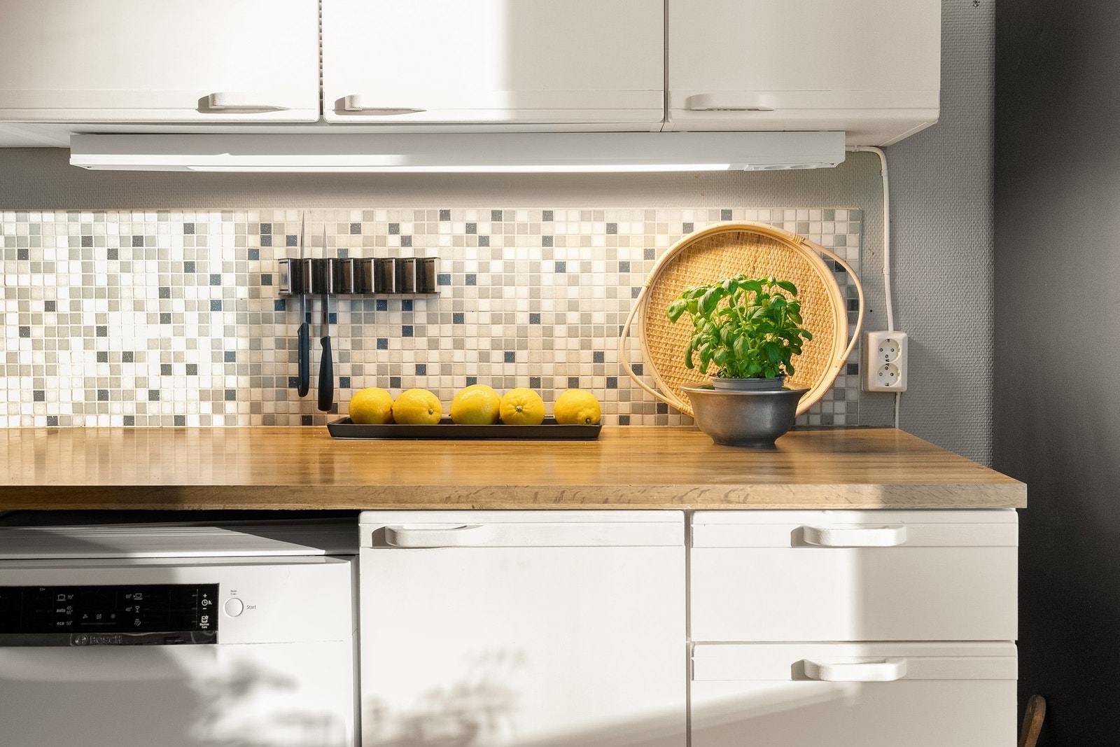 Kjøkkenet har laminat benkeplate med nedfelt kum i rustfritt stål, lysarmatur under overskap og dekorative mosaikkfliser på vegg over kjøkkenbenk. Galleribilde