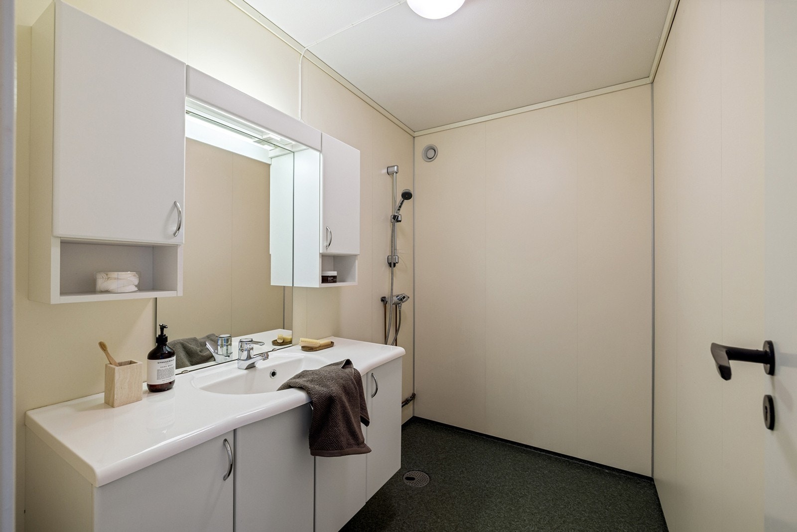 Lyst og romslig bad med gulvbelegg på gulv og baderomsplater på vegger. Innredningen består av et underskap/overskap, servant, speil og armatur med stikk og en dusjsone. Galleribilde