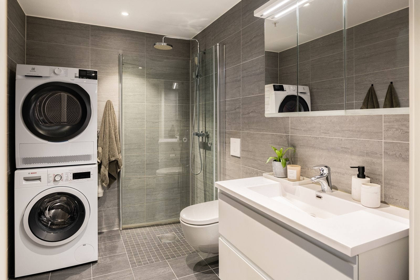 Lekkert baderom med dusjhjørne, vegghengt toalett, servant med underskuffer, overskap med speil og opplegg til vaskemaskin. Galleribilde