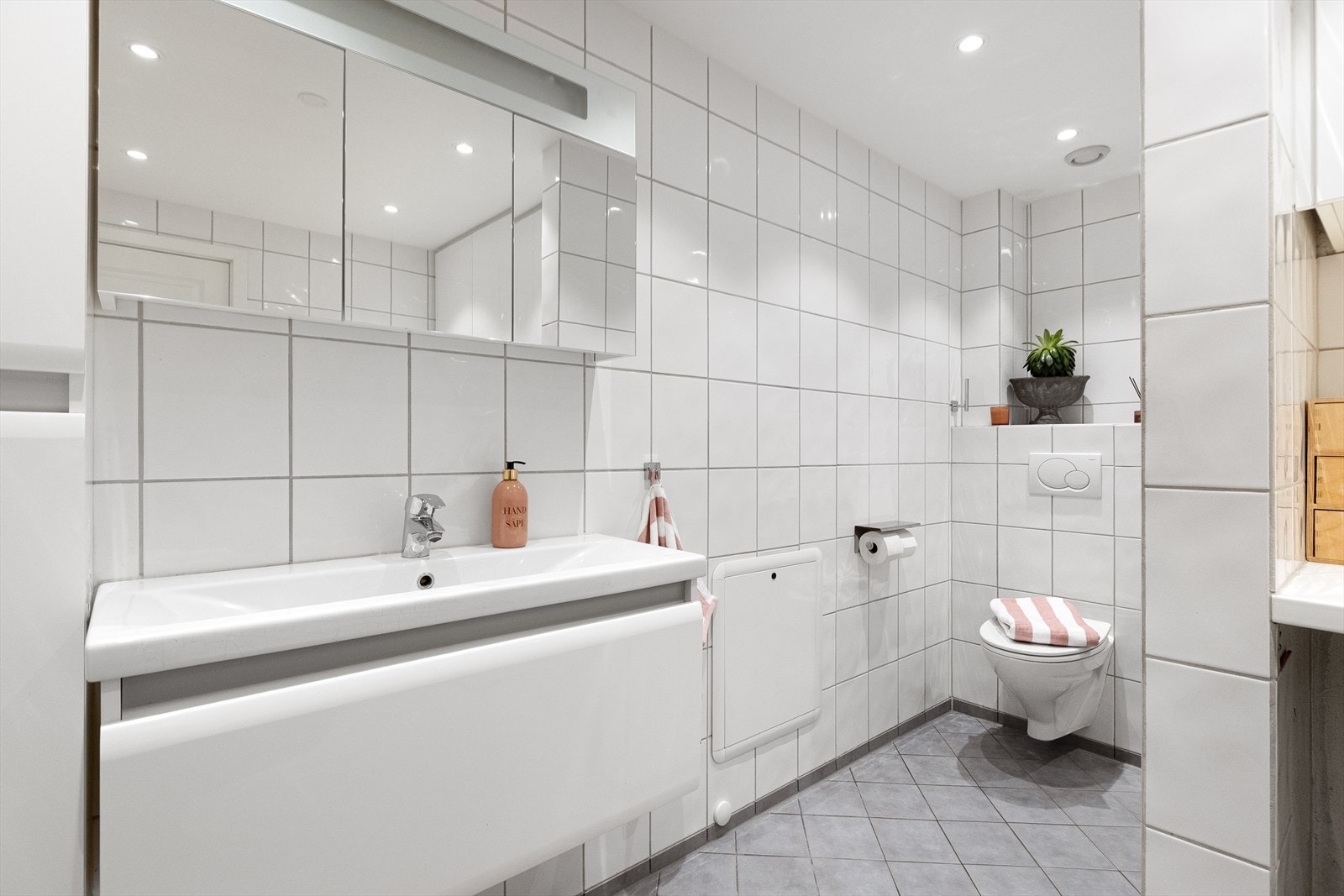 Badet er utstyrt med skap med glatte fronter, veggskap med speil og lys, veggfestet klosett med innebygget sisterne og servant med underskap. Galleribilde