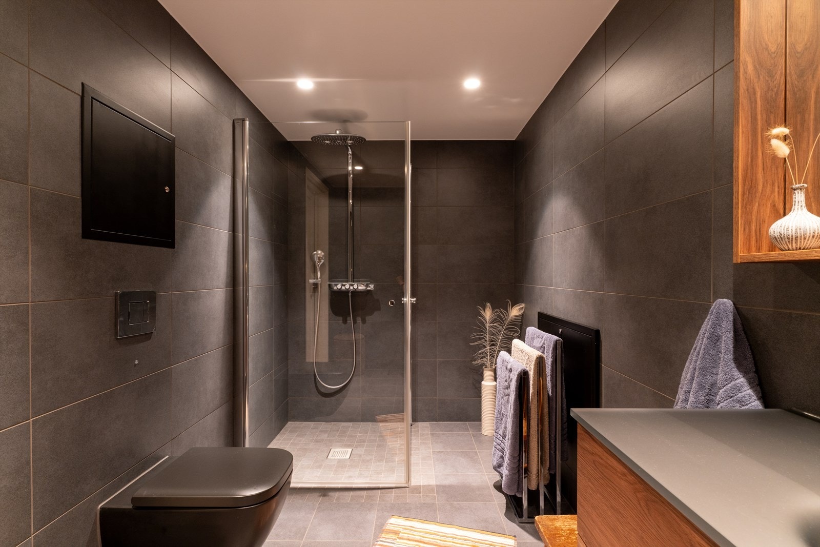 Rommet har flislagte overflater med slitesterke fliser, nedfelt servant i solid innredning, vegghengt toalett med skjult sisterne og dusjhjørne med robuste glassvegger. Elektriske varmekabler gir behagelig varme og økt komfort. Galleribilde