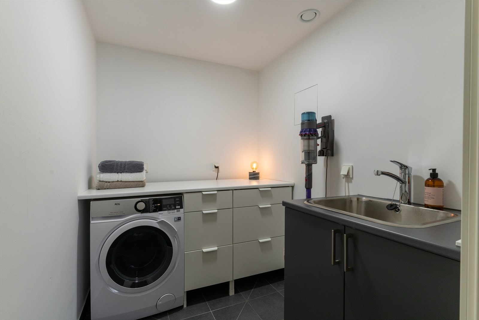 Vaskerommet har gjennomgående praktiske og holdbare materialer. Flislagt gulv med varmekabler, malte plater på vegger og innredning med nedfelt servant. Oppsettet er funksjonelt utformet for å tåle daglig bruk over tid. Galleribilde