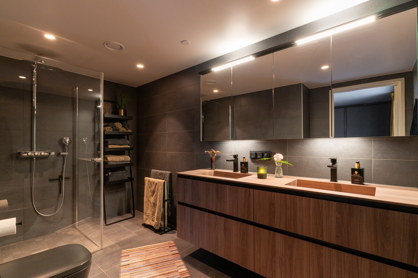 Badet tilknyttet soverom har flislagte overflater med slitesterke fliser, 2 nedfelte servanter i solid innredning, vegghengt toalett med skjult sisterne og dusjhjørne med robuste glassvegger. Elektriske varmekabler gir behagelig varme og økt komfort. Galleribilde