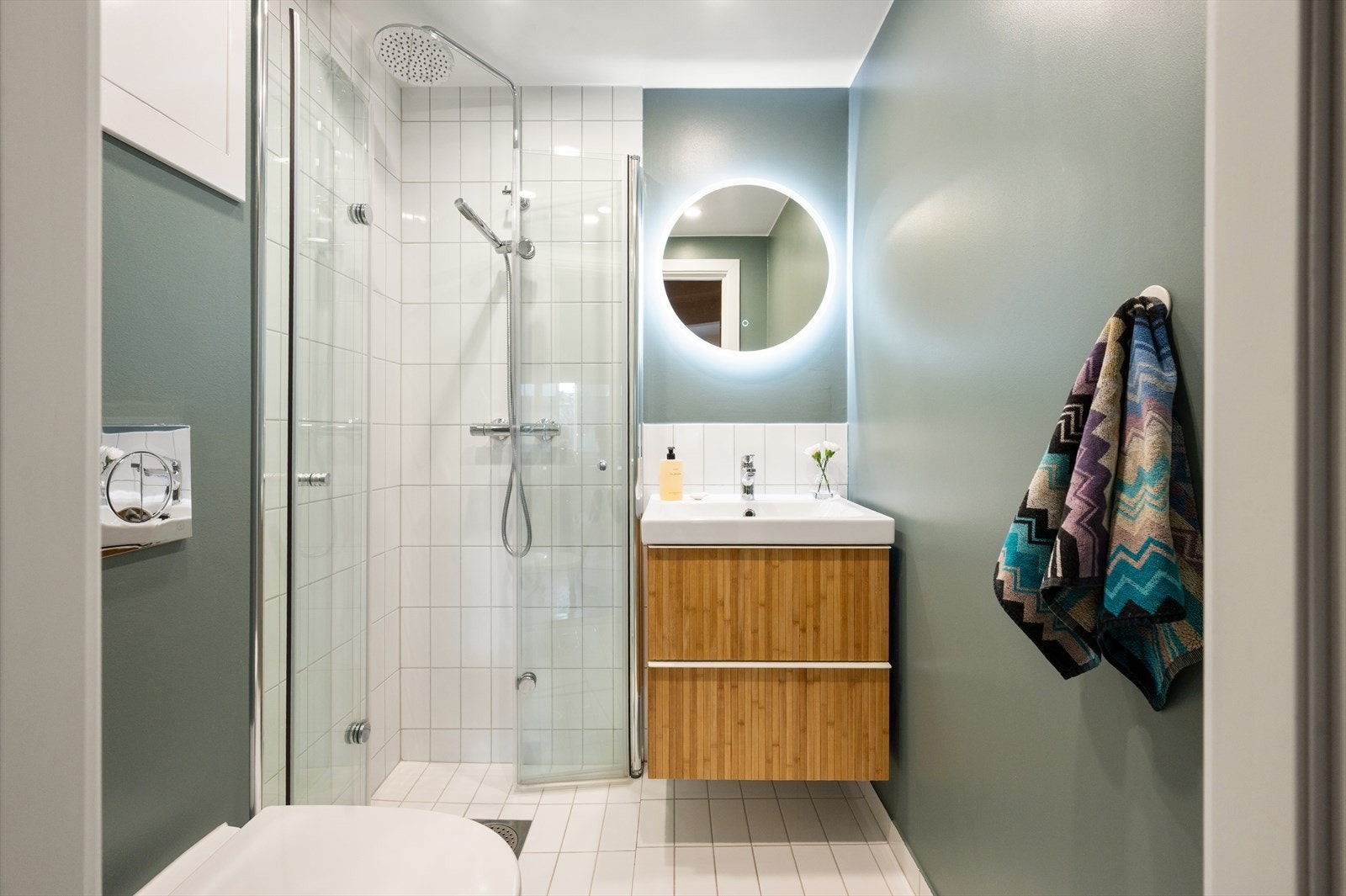 Videre er det innreddet med vegghengt toalett, dusjhjørne med glassdører, samt baderomsinnredning med servant og et moderne rundt speil. Galleribilde