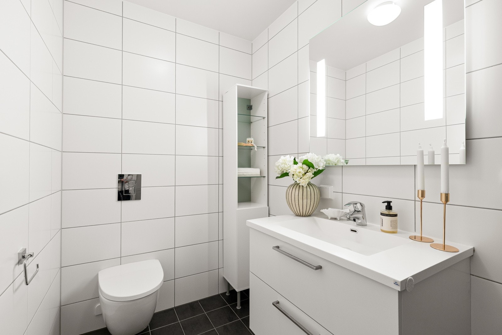 Badet er delikat med mørke gulvfliser og hvite veggfliser, vegghengt toalett, dusjhjørne og hvit baderomsinnredning. Galleribilde