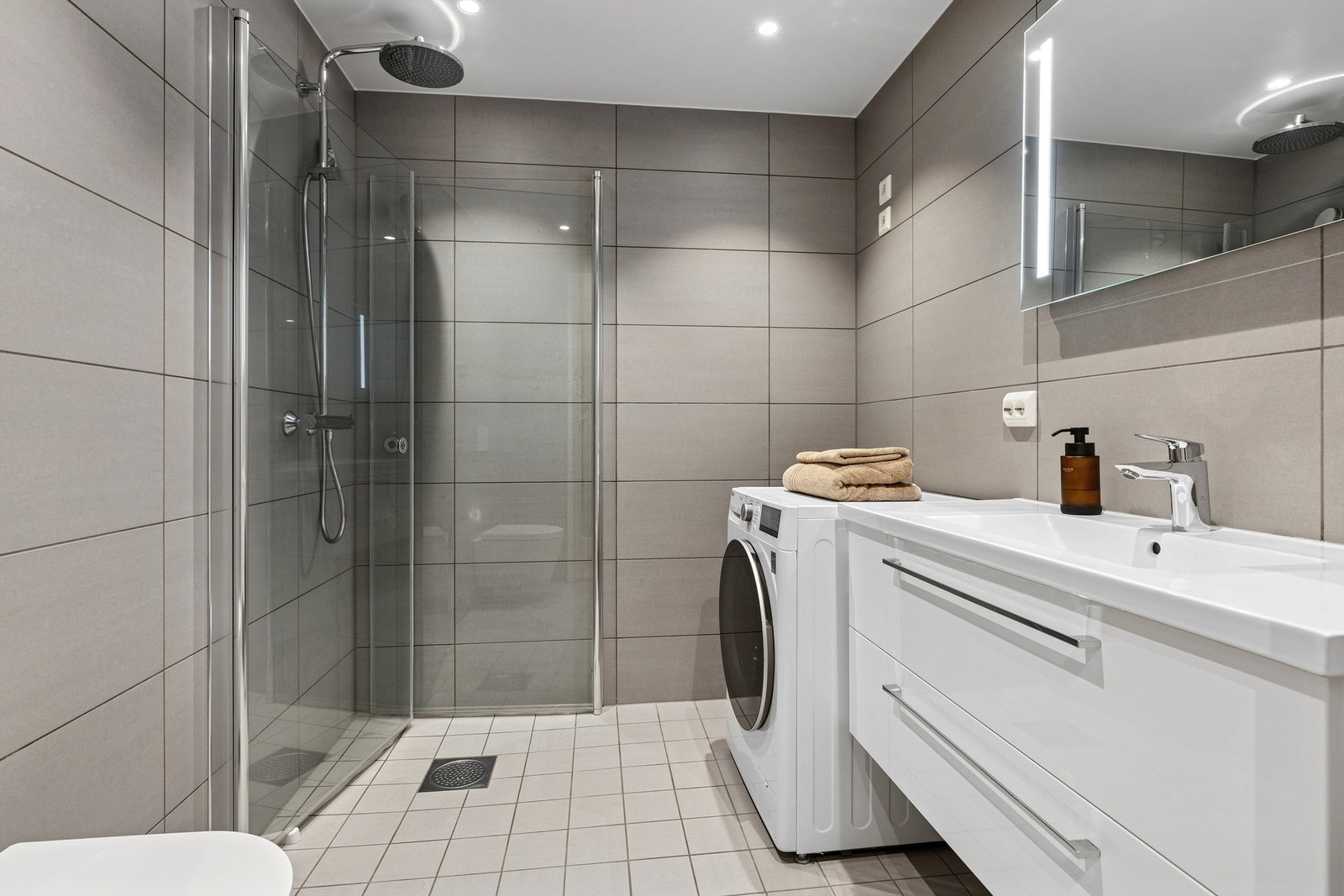 Lekkert flislagt bad med gulvvarme. Her finnes dusjhjørne med innfellbare glassdører, moderne servantinnredning, vegghengt toalett og opplegg for vaskemaskin. Galleribilde