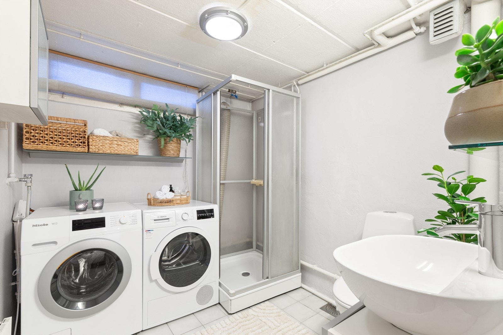 Lyst og romslig vaskerom med opplegg for vaskemaskin, tørketrommel, toalett, servant med innredning og dusjkabinett. Galleribilde
