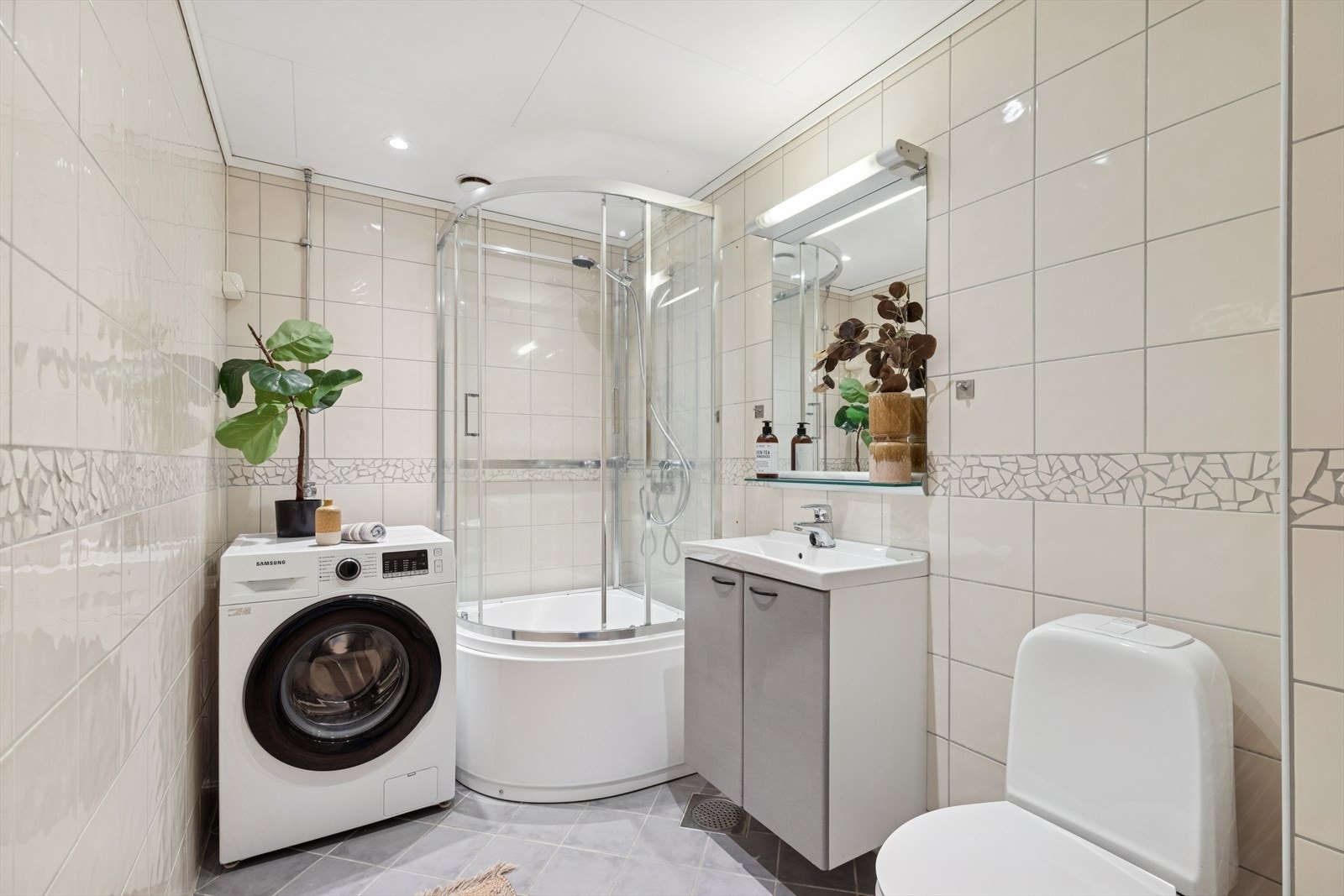 Baderomsinnredningen består av gulvmontert toalett, servant med underskap, speil med belysning og dusjkabinett med skyvedører Galleribilde