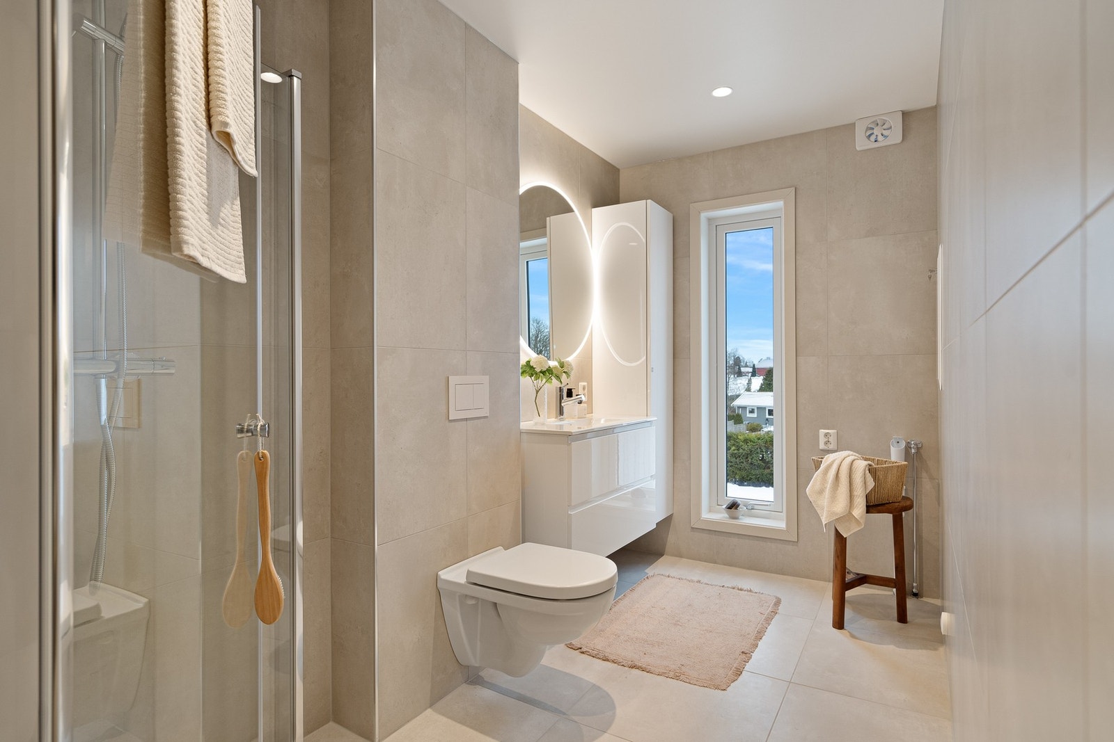 Badet er lekkert og tidsriktig, med store fliser i nøytrale toner, moderne baderomsinnredning og et elegant speil med integrert belysning. Her finner du dusjløsning med glassvegger, vegghengt toalett og servant med oppbevaring. Galleribilde