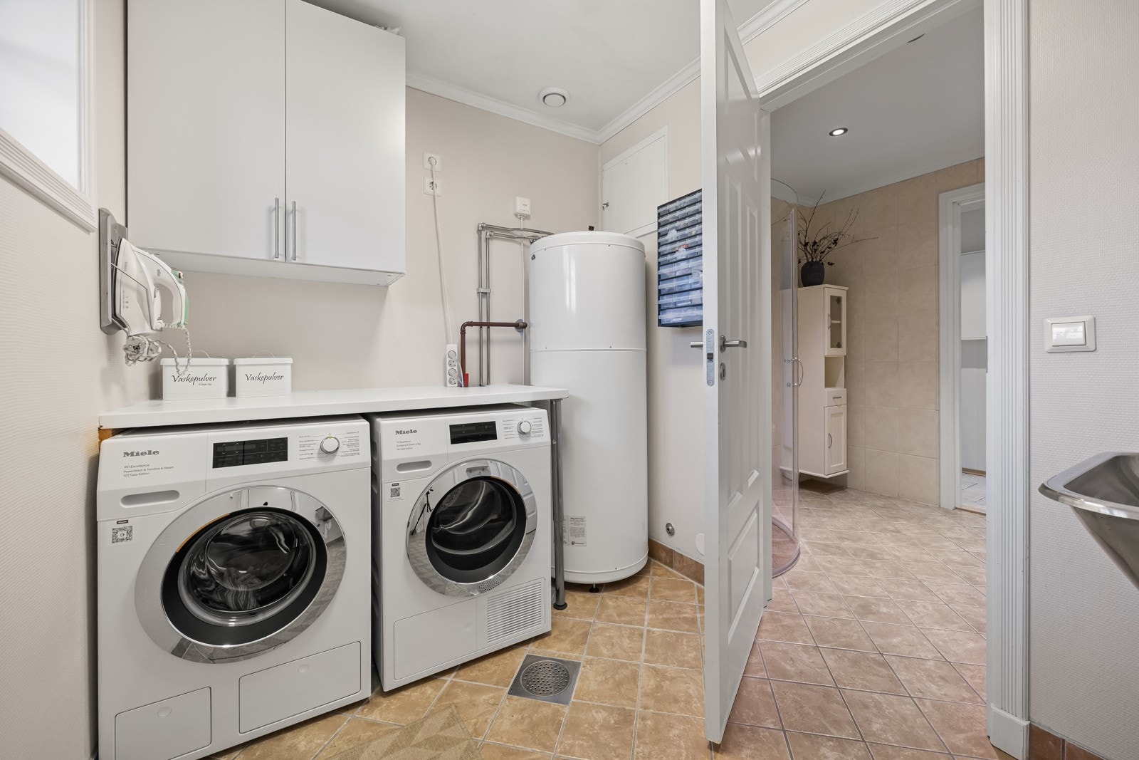 Vaskerommet har malte veggflater og fliser på gulv. Rommet inneholder vegghengte skap, utslagsvask, samt benk med opplegg for vaskemaskin og tørketrommel under. Boligens varmtvannsbereder er plassert her. Galleribilde